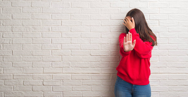 Young brunette woman standing over white brick wall covering eyes with hands and doing stop gesture with sad and fear expression. Embarrassed and negative concept.