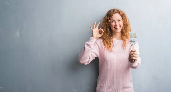 優秀なシンボルの指で サインをしてペイント ブラシを保持している灰色のグランジの壁を越えて若い赤毛の女性 — ストック写真