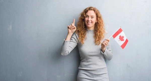 灰色垃圾墙上的年轻红头发的女人持有加拿大国旗惊讶的想法或问题指着一个快乐的脸 — 图库照片