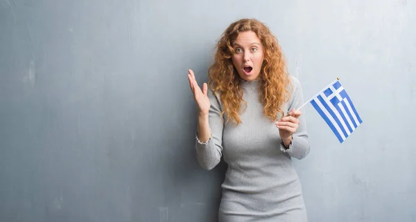 灰色グランジ壁保有上の若い赤毛の女性がギリシャ非常に幸せと興奮 勝者式祝う勝利笑顔で叫び 手を上げたのフラグします — ストック写真