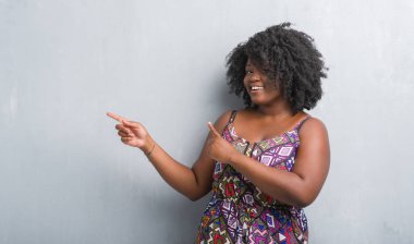 Gri grunge duvar renkli giyen genç Afro-Amerikan kadın elbise gülümseyen ve iki elleri ve parmakları ile tarafına işaret kamera bakıyor.