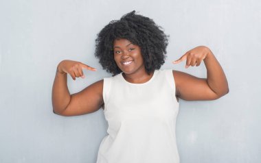 Yüzünde gülümseme ile kendinden emin görünüyorsun, kendini gururlu ve mutlu parmakları ile işaret gri grunge duvarın üzerinden genç Afro-Amerikan kadın.
