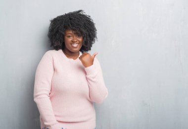 Genç Afro-Amerikan artı boyutu kadın arıyorum ve başparmak yanında yukarıyı mutlu yüz ile gülümseyen kış kazak giyen grunge duvar gri.