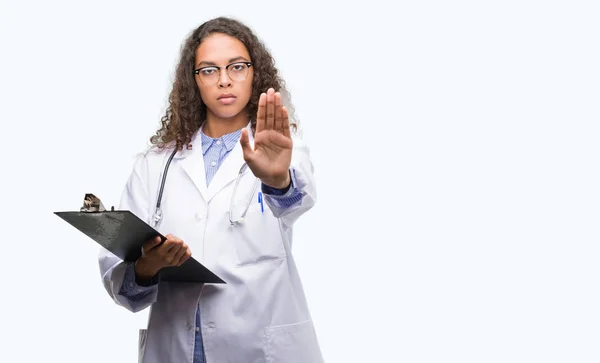 年轻的西班牙裔医生的女人拿着一个剪贴板与张开手做停止标志认真和自信的表达 防御姿态 — 图库照片