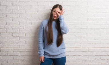 Sürpriz yüz, göz parmaklarının arasından seyir ile genç Chinise kadın Tamam jest yapıyor beyaz tuğla duvarın üzerinden şok. İmansız ifade.