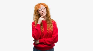 Çene düşünme hakkında soru, dalgın ifade üzerine el ile kırmızı kazak giyen genç Kızıl saçlı kadın. Düşünceli yüzüyle gülümsüyor. Şüphe kavramı.