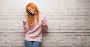 Gülümseyen ve başarı için çığlık tuğla duvar çok mutlu ve heyecanlı kaldırdı, silah ile kazanan jest yapıyor üzerinde duran genç Kızıl saçlı kadın. Kutlama kavramı.