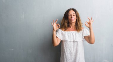 Orta yaş İspanyol gri grunge duvardan duran kadın dinlenmek ve gülümseyen gözlerle parmak ile meditasyon jest yapıyor kapalı. Yoga kavramı.