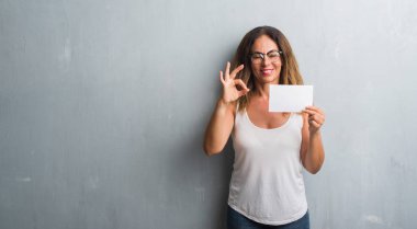 Parmak, mükemmel sembolü ile ok işareti yapıyor boş kartı tutan gri grunge duvarın üzerinden duran orta yaş İspanyol kadın