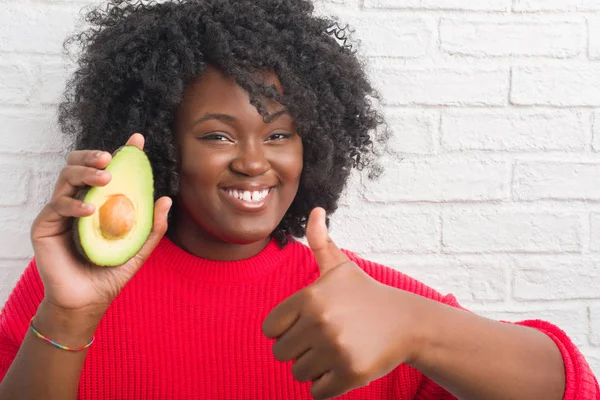 Young african american woman over white brick wall eating avocado happy with big smile doing ok sign, thumb up with fingers, excellent sign