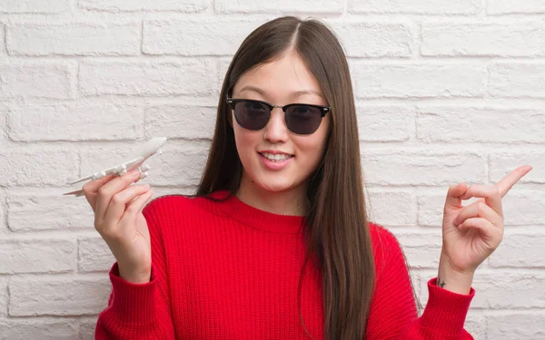 Young Chinese woman over brick wall holding plane toy very happy pointing with hand and finger to the side