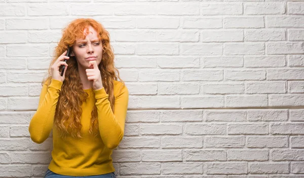 電話深刻な顔考えて質問 非常に混乱しているアイデアについて話をレンガの壁の上に立って若い赤毛の女性 — ストック写真