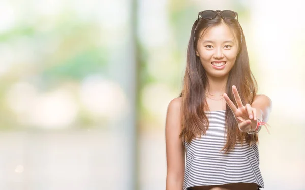 年轻的亚洲妇女戴着太阳镜在孤立的背景下微笑着看着相机 显示手指做胜利标志 第二个 — 图库照片