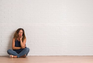 Yerdeki beyaz tuğla duvar düşünme seyir oturan orta yaş İspanyol kadın yorgun ve sıkılmış çapraz silah depresyon sorunları ile.