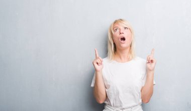 Yetişkin kafkas kadın grunge gri duvarın üzerinden hayran ve yukarıya bakıyor ve parmakları ile işaret ve silah kaldırdı şaşırttı.