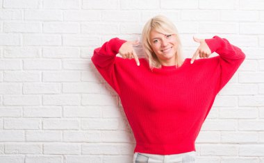 Yüzünde gülümseme ile kendinden emin görünüyorsun, kendini gururlu ve mutlu parmakları ile işaret kış kazak giyen beyaz tuğla duvarın üzerinden yetişkin beyaz kadın.