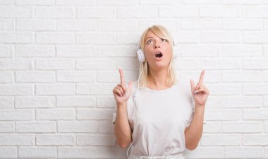 Yetişkin kafkas kadın kulaklık takmış beyaz tuğla duvarın üzerinden hayran ve yukarıya bakıyor ve parmakları ile işaret ve silah kaldırdı şaşırttı.