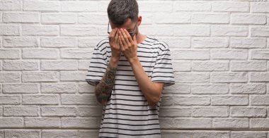 Genç yetişkin adam üzgün ifade yüz eller ile ağlayarak süre kapsayan ile beyaz tuğla duvarı ayakta güneş gözlüğü takıyor. Depresyon kavramı.