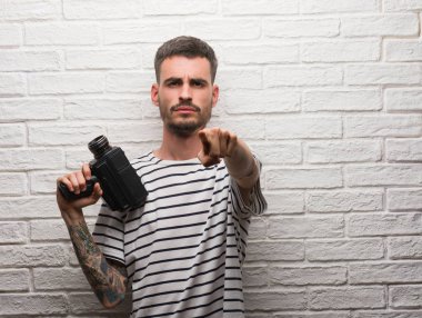 Genç adam filme kamera ve size, parmak ile işaret beyaz tuğla duvarın üzerinden video kamera ayakta tutan el işareti, pozitif ve kendinden emin bir jest ön