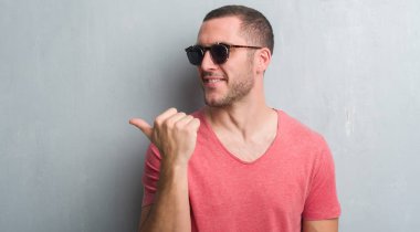 İşaret ve başparmak kadar mutlu yüz gülümseyen tarafı ile gösteren güneş gözlüğü takıyor gri grunge duvarın üzerinden beyaz delikanlı