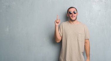 Beyaz delikanlı ile başarılı fikir parmak işaret güneş gözlüğü takıyor gri grunge duvarın üzerinden. Çıktıktan ve mutlu. Bir numara.