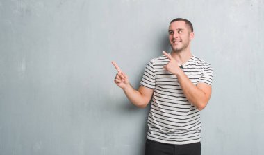 Gülümseyen ve iki elleri ve parmakları ile tarafına işaret kamera bakarak gri grunge duvarın üzerinden beyaz delikanlı.