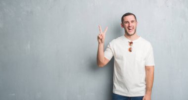 Mutlu yüz zafer işareti yaparken kameraya göz kırpıyor ile gülümseyen güneş gözlüğü takıyor gri grunge duvarın üzerinden Genç beyaz adam. İki numaralı.