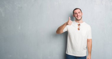 Beyaz delikanlı hareketi el ile mutlu yaşasın yaparken güneş gözlüğü takıyor gri grunge duvarın üzerinden. Kameraya bakarak başarı gösteren ile ifade onaylama.