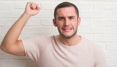 Genç beyaz adam ayakta üzerinde beyaz tuğla duvar rahatsız ve hayal kırıklığına uğramış öfke kavramı ile öfke, çılgın ve yükseltilmiş eliyle diye bağıran bağırarak