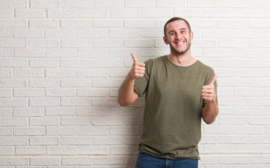Genç beyaz adam üzerinde beyaz tuğla duvar başarı işareti el ile pozitif jest yapıyor ayakta başparmak gülümseyen ve mutlu. Neşeli bir ifadeyle, kazanan jest kameraya bakıyor.