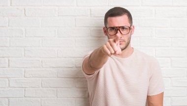 Genç beyaz adam ayakta üzerinde beyaz tuğla duvar gözlük kamera ve size parmak ile işaret, el işareti, pozitif ve kendinden emin bir jest ön