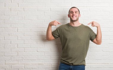 Yüzünde gülümseme ile kendinden emin görünüyorsun, kendini gururlu ve mutlu parmakları ile işaret beyaz tuğla duvarın üzerinden ayakta beyaz delikanlı.