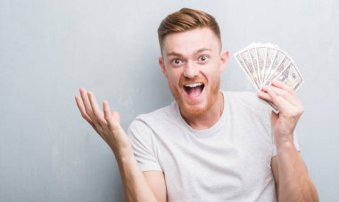 Genç Kızıl saçlı adam dolar çok mutlu ve heyecanlı, kazanan ifade ile çığlık zaferini kutluyor büyük holding gri grunge duvarın üzerinden gülümse ve el kaldırdı
