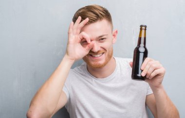 Genç Kızıl saçlı adam bira şişesi ile mutlu yüz el parmaklarının arasından seyir göz üzerinde ok işaretiyle yapıyor gülümseyen içme gri grunge duvarın üzerinden