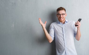 Genç Kızıl saçlı adam gri grunge duvar büyük gülümseme ile çığlık zaferini kutluyor ve ellerini kaldırdı smartphone çok mutlu ve heyecanlı, kazanan ifade kullanarak gönderme mesaj