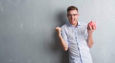 Young redhead man over grey grunge wall holding piggy bank screaming proud and celebrating victory and success very excited, cheering emotion