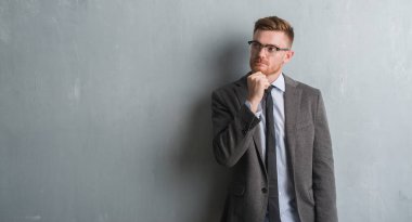 Genç redhead zarif iş adamı gri grunge duvar ciddi bir yüz düşünme hakkında soru, çok karışık bir fikir üzerinde