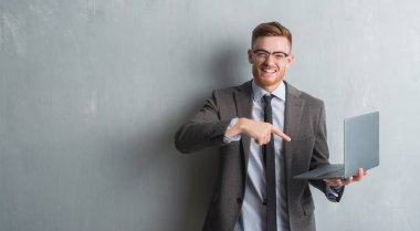 Genç redhead zarif iş adamı dizüstü el ve parmak ile işaret çok mutlu kullanarak gri grunge duvar üzerinde