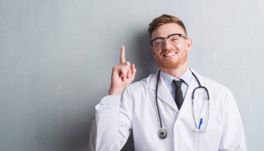 Genç redhead Doktor gri grunge duvarın bir fikirle sürpriz üzerinden adamım ya da gülen yüz, bir numara ile işaret parmağı soru