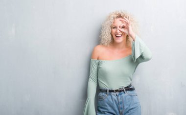 Grunge üzerinde Kıvırcık saçlı sarışın genç kadın gülümsüyor, parmakları ile mutlu yüz üzerinden seyir göz el ile ok jest yapıyor arka plan gri.