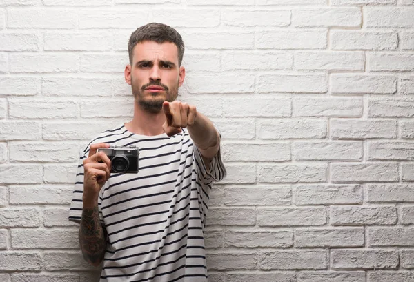 Young man holding vintage camera standing over white brick wall pointing with finger to the camera and to you, hand sign, positive and confident gesture from the front