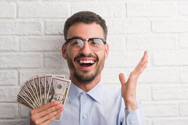 Young hipster business man holding dollars very happy and excited, winner expression celebrating victory screaming with big smile and raised hands