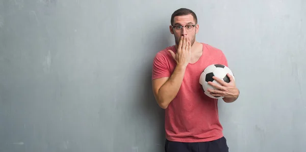 Young caucasian man over grey grunge wall holding soccer football ball cover mouth with hand shocked with shame for mistake, expression of fear, scared in silence, secret concept