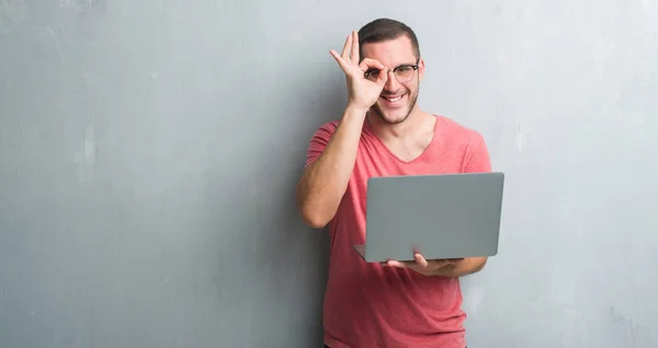 年轻的白种人在灰色的垃圾墙使用电脑笔记本电脑与快乐的脸微笑做 标志与手在眼睛看通过手指 — 图库照片