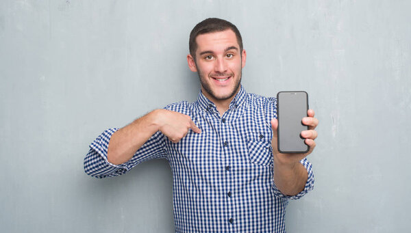 Молодой кавказский мужчина над серой гранж-стеной показывает пустой экран смартфона с неожиданным лицом, указывающим пальцем на себя
