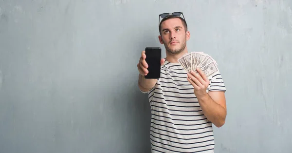 Young Caucasian Man Grey Grunge Wall Holding Dollars Smarphone — Stock Photo, Image