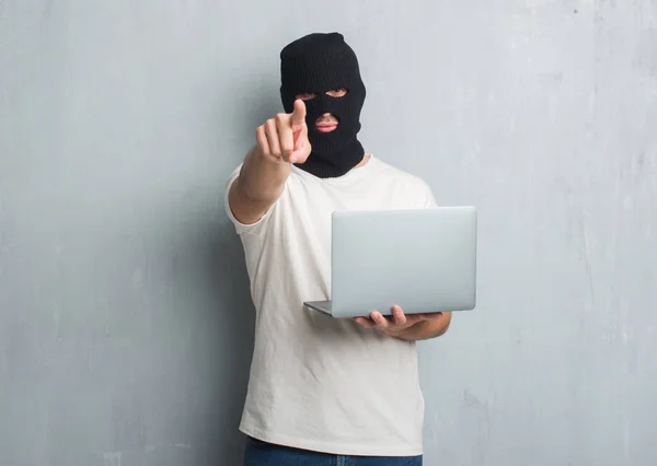 年轻的白种人黑客在灰色的垃圾墙做网络攻击使用笔记本电脑指向相机和你 正面和自信的手势从前面 — 图库照片