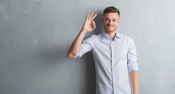 Young redhead business man over grey grunge wall smiling positive doing ok sign with hand and fingers. Successful expression.