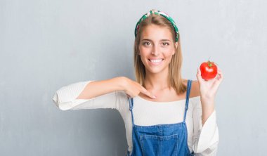 Güzel genç kadın kendine sürpriz yüz işaret parmağı ile taze domates yeme grunge gri duvarın üzerinden