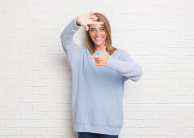 Beyaz tuğla duvar elle yapma çerçeve ve parmakları ile mutlu yüz gülümseyen güzel genç kadın. Yaratıcılık ve Fotoğrafçılık kavramı.